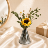 A small clear glass vase holding a sunflower and greenery stems, placed on draped cream fabric with a warm neutral background and a gift box in the side.