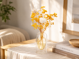 A clear glass vase filled with yellow dried flowers and a white decorative element, placed on a light wood table in a softly lit room with books and a framed print in the background.