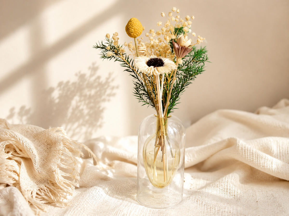A small clear glass vase holding a dried flower arrangement with cream baby's breath, a yellow pom-pom style bloom, green sprigs, and a white daisy with a dark center, styled on a cream fabric surface with a fringed textile and warm sunlight shadows.