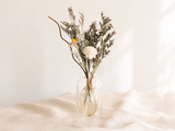 Dried flowers arranged in a clear glass vase on a draped cream fabric surface against a soft white background.