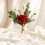 A floral arrangement featuring vibrant red flowers, green pine branches, and dried beige grasses in a clear glass vase with a gold-toned rim, set against a draped cream fabric background.