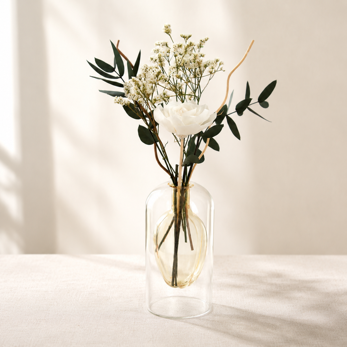 A clear glass vase with a double-walled design holding a dried floral arrangement featuring a white rose, white baby's breath, and green eucalyptus leaves, set on a light beige fabric surface against a warm beige wall with soft window shadows.
