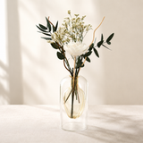 A clear glass vase with a double-walled design holding a dried floral arrangement featuring a white rose, white baby's breath, and green eucalyptus leaves, set on a light beige fabric surface against a warm beige wall with soft window shadows.