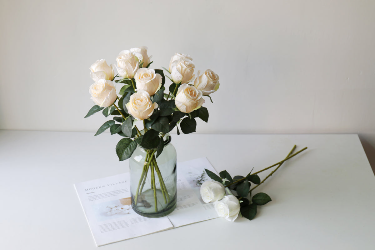 Cream faux roses in a green glass jar with two white stems laid beside