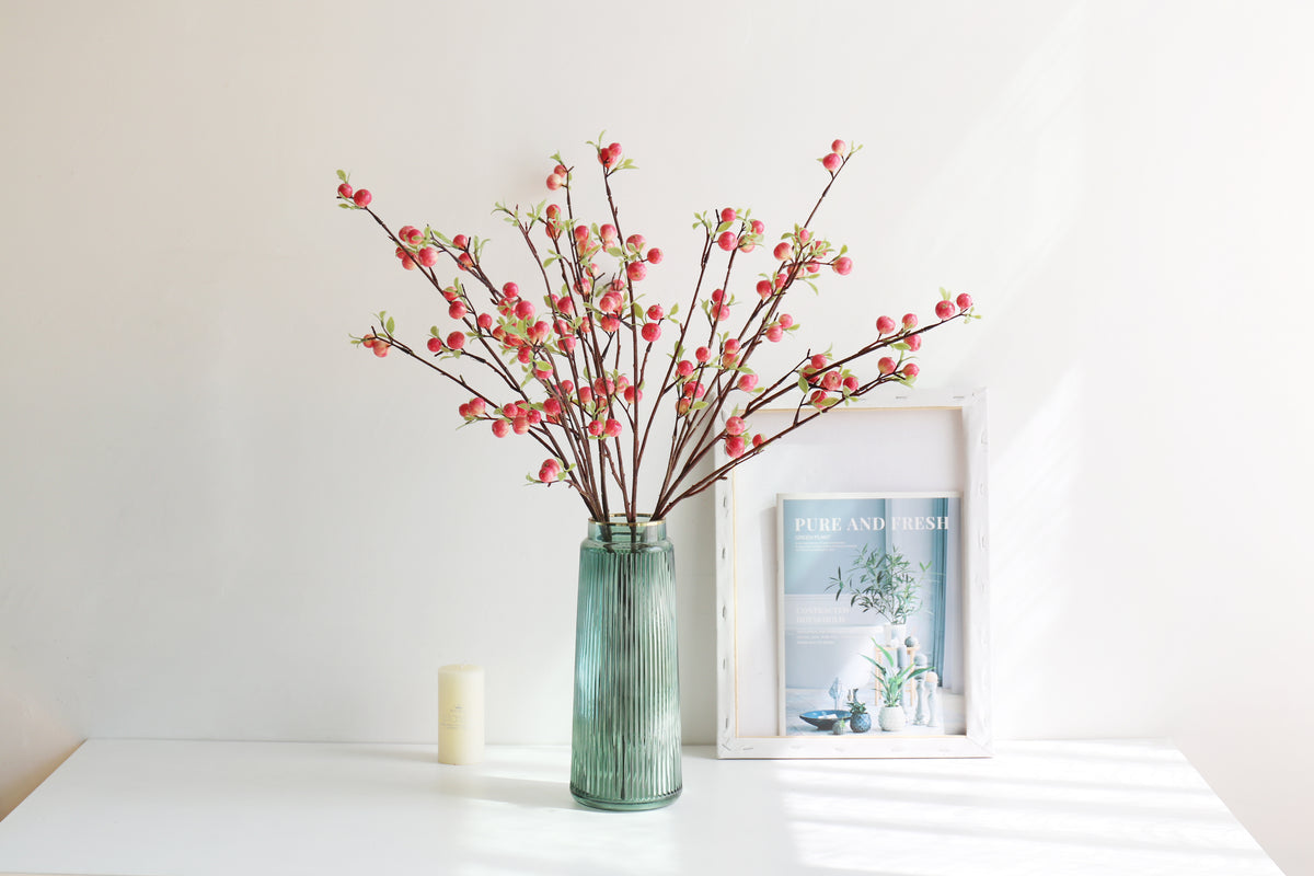 Coral red faux berry branches with slender green leaves in a green glass vase