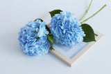 Blue faux hydrangea stems with green leaves, laid flat on a wooden frame