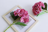 purple faux hydrangea stems with green leaves, laid flat on a wooden frame