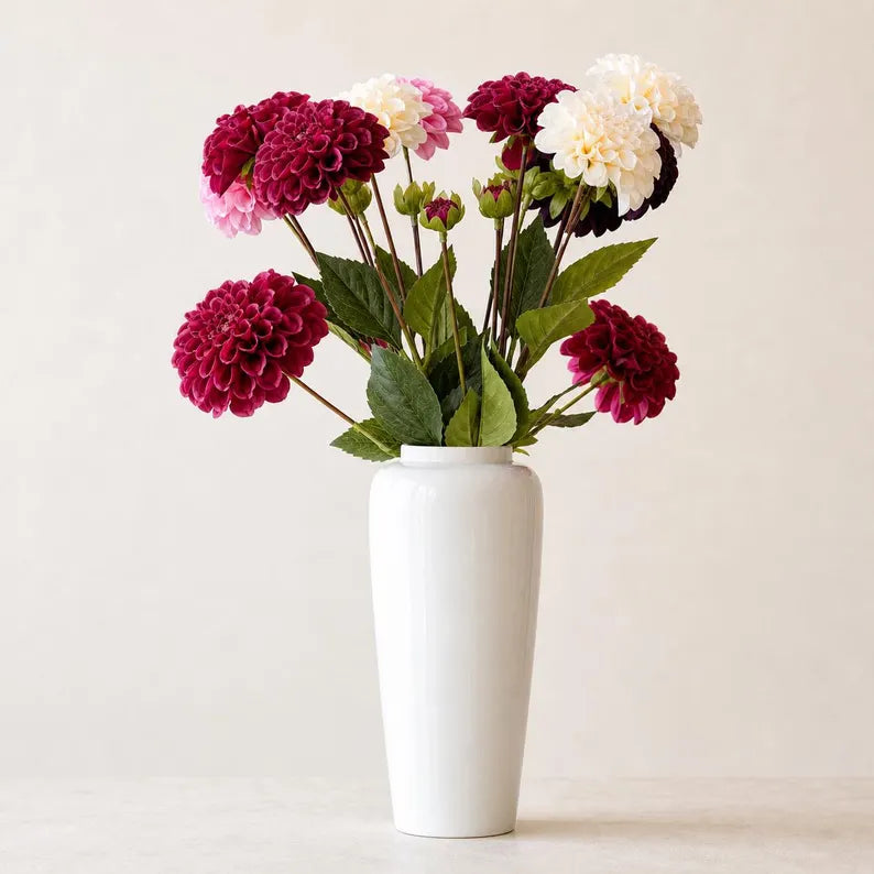 Deep red and cream faux dahlia stems with green foliage in a tall white vase