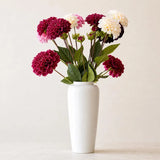Deep red and cream faux dahlia stems with green foliage in a tall white vase