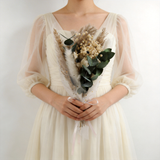 A woman wearing a cream tulle dress with sheer puff sleeves holds a dried floral bouquet with pampas grass, baby's breath, and green leaves against a plain light background.